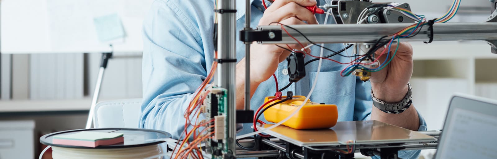 Person using a multimeter with a 3D printer, surrounded by wires and filament spools.