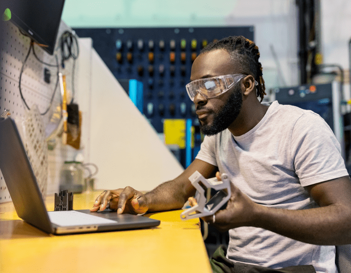 A person wearing safety goggles works on a laptop while holding a mechanical component in a workshop setting.