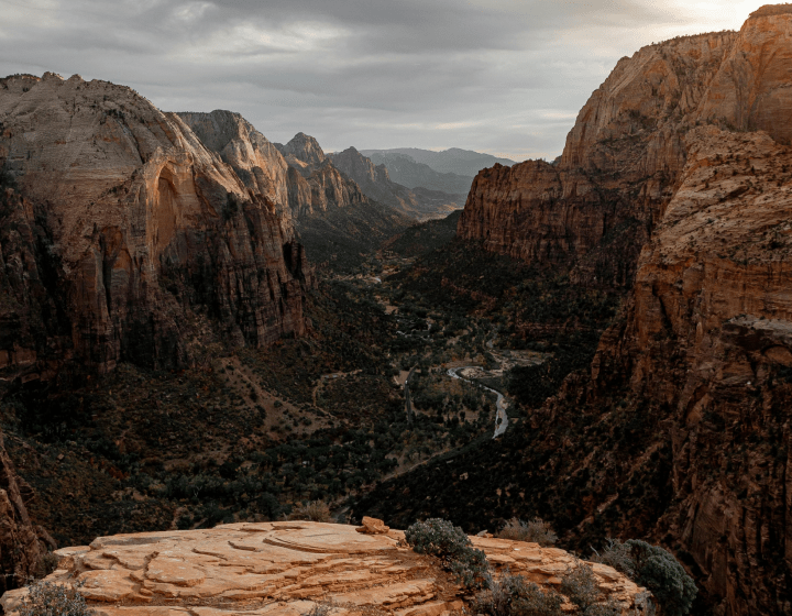 A canyon landscape with towering rock formations, a river winding through the canyon floor, and a rocky ledge in the foreground.