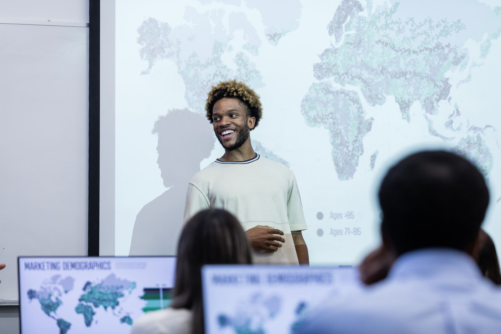 Young African American man standing in front of a class, smiling to someone out of view. Behind him is a projector screen showing a map of the world.