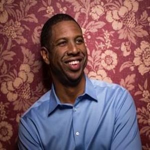 Hakeem Oluseyi smiling in front of a red floral wallpaper, wearing a light blue dress shirt