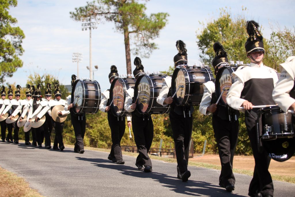 TJC Apache Band marching in formation with drumline and cymbal players during a daytime tradition.