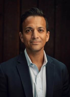 Vin Gupta in a navy blazer and white shirt, posing against a dark wood background.