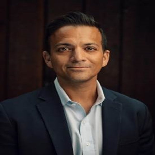 Vin Gupta in a navy blazer and white shirt, posing against a dark wood background.