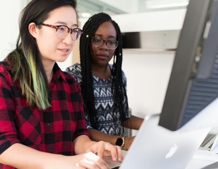 Two women wearing glasses collaborate on a computer, focused on their work in a bright, modern office setting.