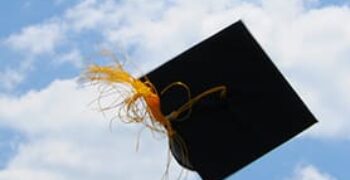 Graduation cap with yellow tassel in the air against a blue sky with clouds.