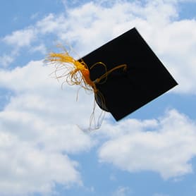 Graduation cap with yellow tassel in the air against a blue sky with clouds.