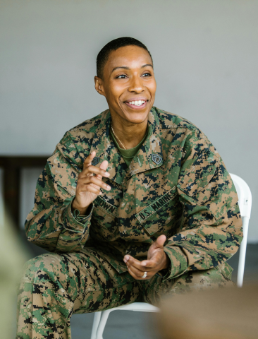Person in U.S. Marines camo uniform, smiling and gesturing while seated on a white chair.